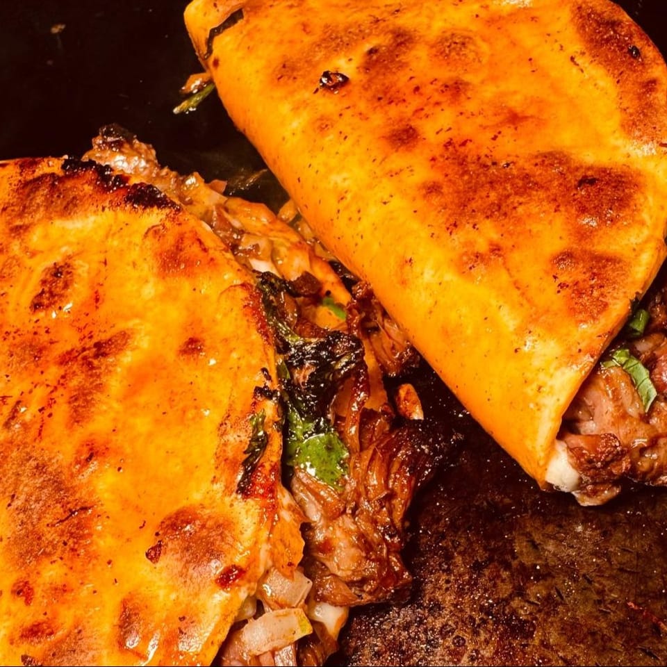 Two golden-brown birria tacos, filled with meat and cilantro, cooking on a dark griddle.